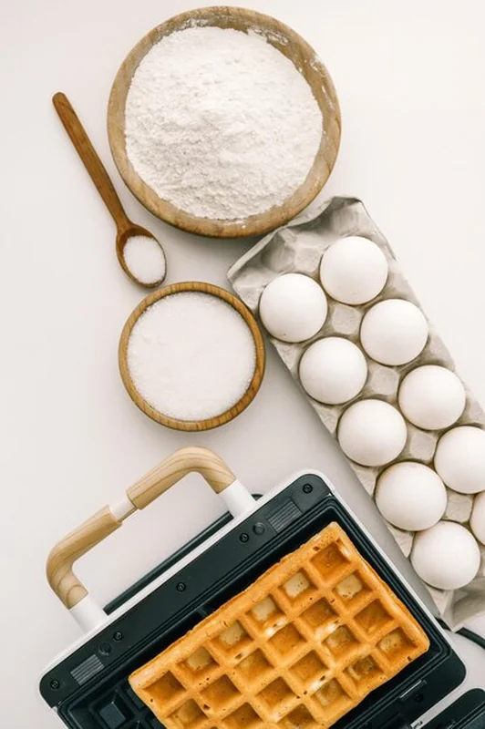 Składniki do gofrów na tle maszyny do waflow - mąka, jajka, cukier, złoty wafelek